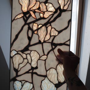 May include: A stained glass window panel featuring a floral design with white and beige glass pieces. The flowers are arranged in a symmetrical pattern with a black metal frame.