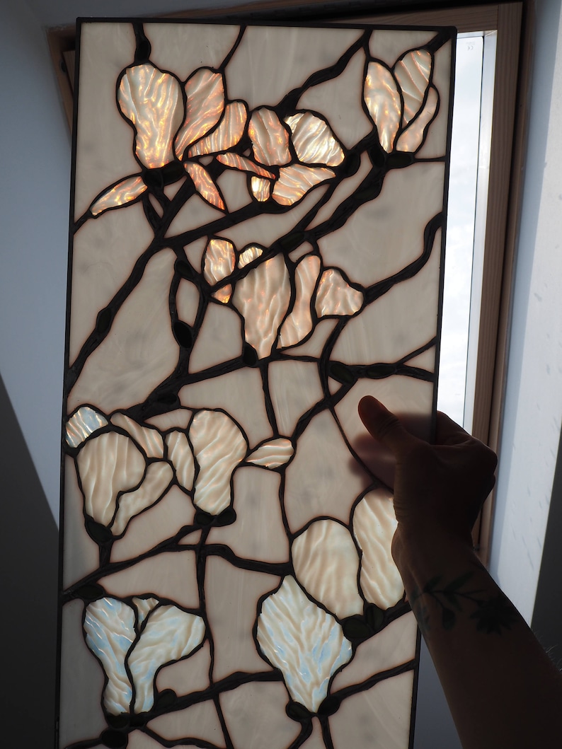 May include: A stained glass window panel featuring a floral design with white and beige glass pieces. The flowers are arranged in a symmetrical pattern with a black metal frame.