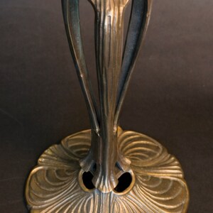 May include: A bronze lamp base with an intricate, floral design. The base has a wide, circular foot with a raised center and a slender, ornate stem.