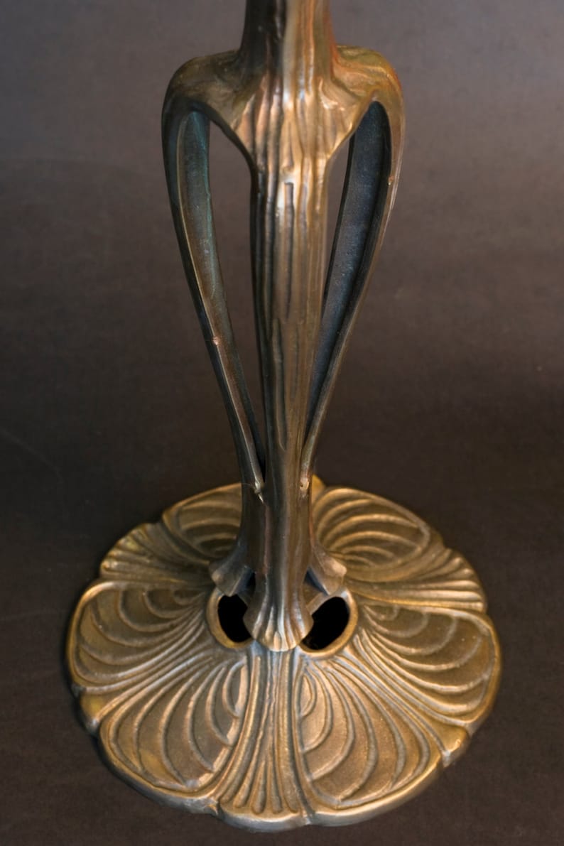 May include: A bronze lamp base with an intricate, floral design. The base has a wide, circular foot with a raised center and a slender, ornate stem.