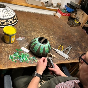 May include: A person working on a mosaic lampshade. The lampshade is green and black with a geometric pattern. The person is using a pair of pliers to cut small pieces of glass.
