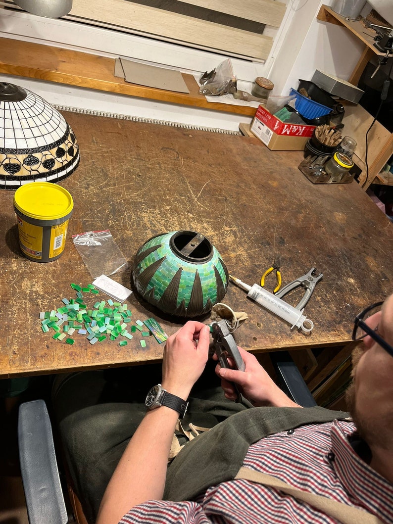 May include: A person working on a mosaic lampshade. The lampshade is green and black with a geometric pattern. The person is using a pair of pliers to cut small pieces of glass.