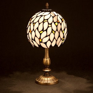 K&ouml;nnte beinhalten: Eine Tischlampe aus wei&szlig;em und goldenem Buntglas mit einem Blattmotiv. Die Lampe hat einen runden Schirm und einen bronzenen Sockel.
