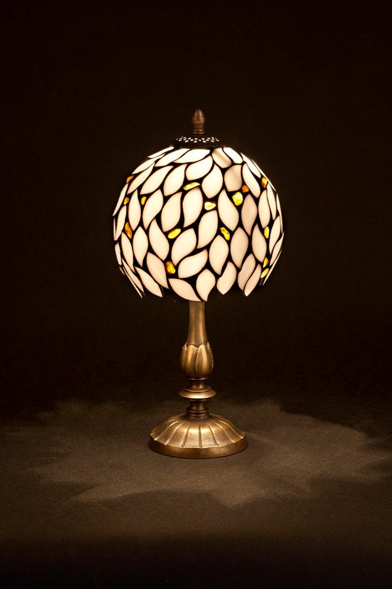 K&ouml;nnte beinhalten: Eine Tischlampe aus wei&szlig;em und goldenem Buntglas mit einem Blattmotiv. Die Lampe hat einen runden Schirm und einen bronzenen Sockel.