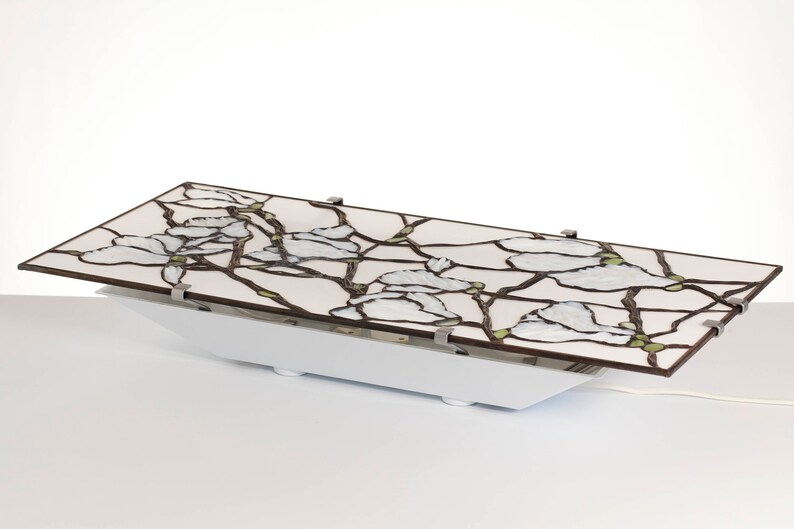 May include: A white ceiling light fixture with a stained glass shade featuring a floral design of white blossoms and brown branches. The shade is attached to a white base with silver clips.