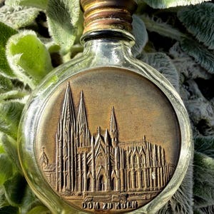 Pode incluir: Uma garrafa de vidro transparente com uma tampa de metal cor de bronze e um emblema redondo cor de bronze. O emblema apresenta um relevo detalhado de uma catedral com o texto "DOM ZU KOLN" na parte inferior. A garrafa é cercada por folhagem verde.