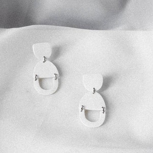 May include: A pair of white geometric earrings with a semi-circle and oval shape. The earrings are made of a smooth, matt material.