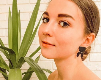 Puede incluir: Una mujer con cabello castaño y un piercing en la nariz lleva pendientes negros con forma de oso. Está mirando hacia un lado y sonriendo.