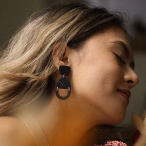 May include: A woman with long brown hair wearing black geometric earrings. She is wearing a white tank top and has a bouquet of pink flowers in her hand.