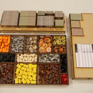 May include: A wooden organizer for the board game "The Crew: The Quest for Planet Nine". The organizer has compartments for the game's cards, tokens, and meeples. The organizer is made of light brown wood and has labels for each compartment.