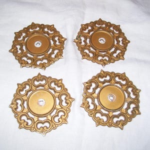 May include: Four gold-colored, ornate decorative medallions. Each medallion has a central circular element with a clear, faceted gem and is surrounded by a detailed, openwork design. The medallions are arranged on a white surface.