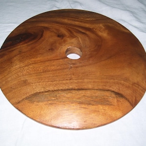 May include: A circular wooden disc with a smooth, polished finish and a prominent natural grain pattern in shades of medium to dark brown. The object features a single, centered circular hole, creating a simple, flat, and symmetrical design.
