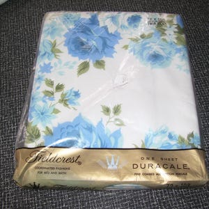 Puede incluir: Una s&aacute;bana plana individual empaquetada con un estampado floral de rosas azules y hojas verdes sobre un fondo blanco. La s&aacute;bana est&aacute; etiquetada como "Fieldcrest Duracale" y mide 183 x 274 cm.