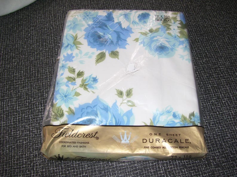 Puede incluir: Una s&aacute;bana plana individual empaquetada con un estampado floral de rosas azules y hojas verdes sobre un fondo blanco. La s&aacute;bana est&aacute; etiquetada como "Fieldcrest Duracale" y mide 183 x 274 cm.
