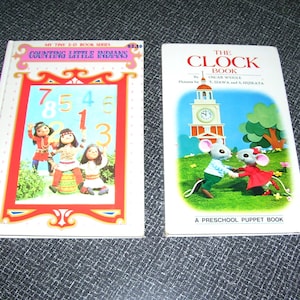May include: Two vintage children's books. One book titled "Counting Little Indians" features illustrations of children and numbers. The other book, "The Clock Book," shows a clock tower and cartoon mice. Both books are on a dark surface.