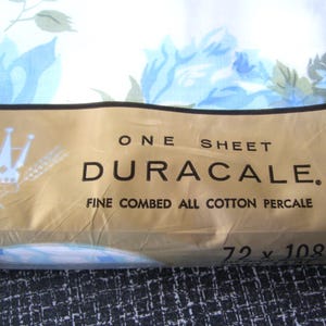 Puede incluir: Un paquete de s&aacute;bana Duracale de percal de algod&oacute;n peinado fino. La s&aacute;bana es blanca con un estampado floral azul. El paquete es de color beige con el texto "ONE SHEET DURACALE" y mide 183 x 274 cm.