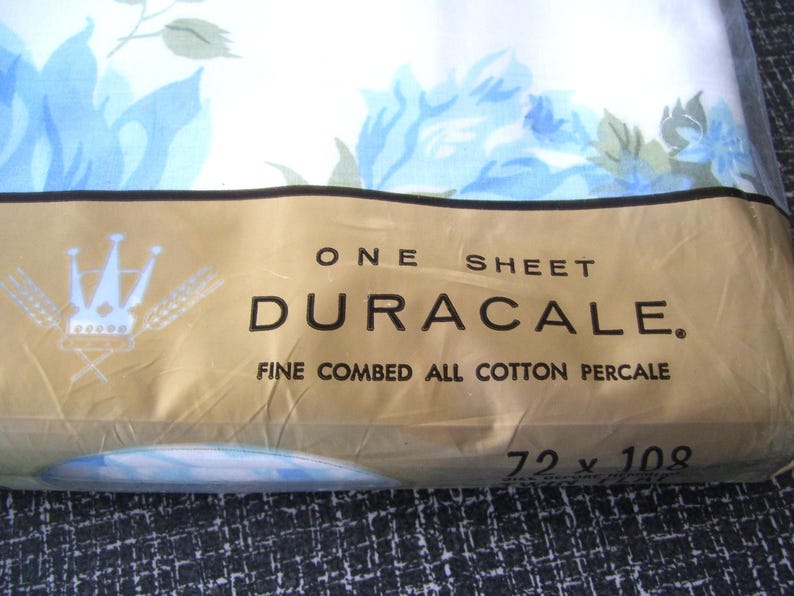 Puede incluir: Un paquete de s&aacute;bana Duracale de percal de algod&oacute;n peinado fino. La s&aacute;bana es blanca con un estampado floral azul. El paquete es de color beige con el texto "ONE SHEET DURACALE" y mide 183 x 274 cm.