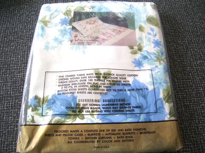 Puede incluir: Un juego de s&aacute;banas Fieldcrest empaquetadas. El embalaje es de pl&aacute;stico transparente, revelando un vistazo al patr&oacute;n floral en el interior. Las s&aacute;banas presentan un fondo blanco con dise&ntilde;os florales azules y verdes. El texto en el embalaje proporciona detalles.