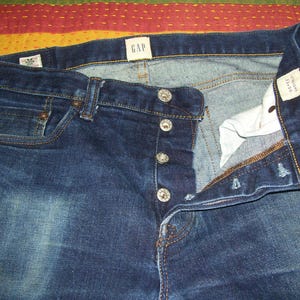 May include: Dark blue denim jeans with a button fly. The jeans have a faded wash and feature a small red accent on the front pocket. The GAP label is visible at the waistband. The jeans are straight leg.