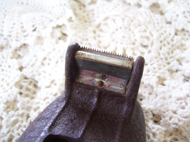 May include: Close-up of a vintage brown tape dispenser. The dispenser has a serrated blade for cutting tape and a textured surface. The image is set against a white, lace-like background.