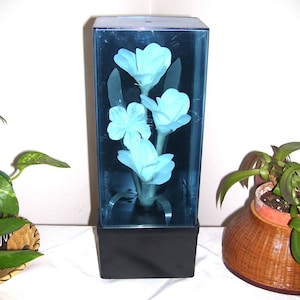 May include: A rectangular black base supports a tall, transparent blue acrylic case containing three light blue artificial flowers with green stems and leaves. The flowers are arranged vertically inside the enclosure, which is sealed at the top.