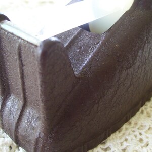 May include: A dark brown tape dispenser with a textured surface. The dispenser has a classic design with a blade for cutting tape. The tape is visible in the dispenser.