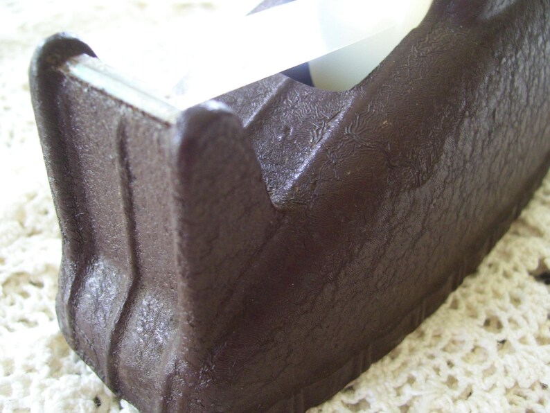 May include: A dark brown tape dispenser with a textured surface. The dispenser has a classic design with a blade for cutting tape. The tape is visible in the dispenser.