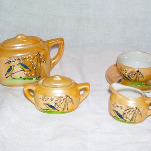 May include: A vintage miniature tea set with a teapot, sugar bowl, creamer, teacup, and saucer. The set is a light orange color with a pearlescent finish. Each piece is decorated with a painted design of birds and foliage.