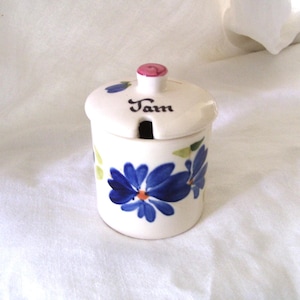 May include: A white ceramic jam pot with a matching lid featuring a small pink knob. The pot is decorated with hand-painted blue flowers and green leaves. The word Jam is written in black script on the lid, which includes a small notch for a spoon.