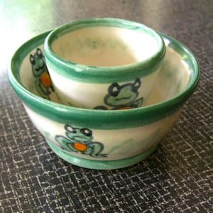 May include: A set of two ceramic bowls, one nested inside the other. The bowls are white with a green rim and feature hand-painted frog illustrations in green and orange. The bowls are suitable for serving food.