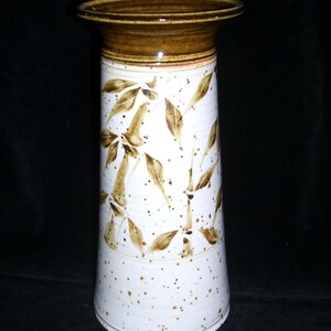 May include: A tall, ceramic vase with a wide, brown rim and a cylindrical body. The vase is white with brown bamboo leaf designs and speckled accents. The vase is approximately 10 inches tall.
