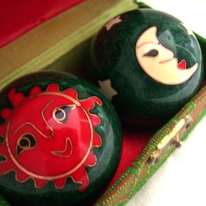 May include: Two green Chinese health balls with red and gold sun and moon designs, resting in a red and green lined box. The sun has a smiling face, and the moon has a serene expression. The box is green with a gold clasp.