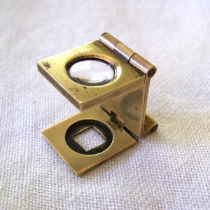 May include: A brass folding linen tester magnifying glass with a square viewing aperture and a circular lens. The metal frame features a hinged design and the word FRANCE is stamped clearly on the base plate near the hinge mechanism.