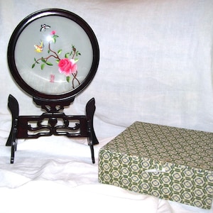 May include: A round, dark wood framed decorative piece with a floral and butterfly design. The piece is displayed on a matching stand. A patterned box is next to the stand.