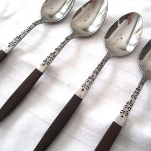 May include: Four spoons with stainless steel bowls and dark brown handles. The handles are a dark brown color and the stems are decorated with a black floral design. The spoons are arranged diagonally on a white surface.