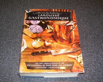 Puede incluir: Un libro de tapa dura titulado Larousse Gastronomique, The World Authority. La portada muestra una olla de cobre, un cuchillo de chef y col lombarda cortada sobre una superficie de madera. El texto incluye The First American Edition of the Encyclopedia of Food, Wine and Cookery, 8500 Recipes, 1000 Illustrations.