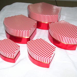 May include: A set of five hexagonal gift boxes in varying sizes, each featuring a red and white diagonal striped lid and a solid metallic red base. The boxes are arranged in a cluster, showcasing their glossy finish and consistent design.