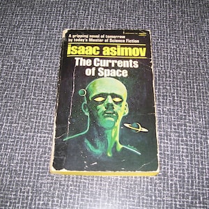 May include: A vintage paperback book titled "The Currents of Space" by Isaac Asimov. The cover features a green-skinned alien with glowing yellow eyes against a dark background. The book's title and author's name are in bold, yellow and white text.