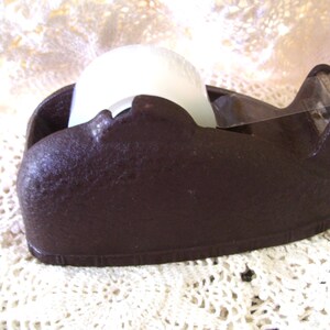 May include: A dark brown tape dispenser with a textured surface. The dispenser holds a roll of white tape and has a clear plastic cutting edge. The dispenser is sitting on a white lace surface.