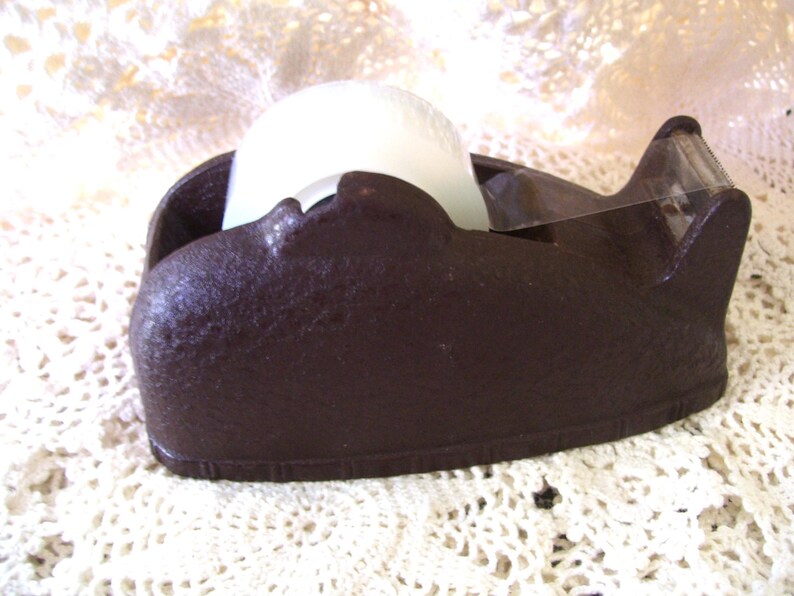 May include: A dark brown tape dispenser with a textured surface. The dispenser holds a roll of white tape and has a clear plastic cutting edge. The dispenser is sitting on a white lace surface.