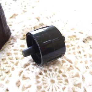 May include: A black plastic bottle stopper with a cylindrical body and a small, protruding peg. The stopper is set against a white, crocheted background. The stopper is a dark, solid color.