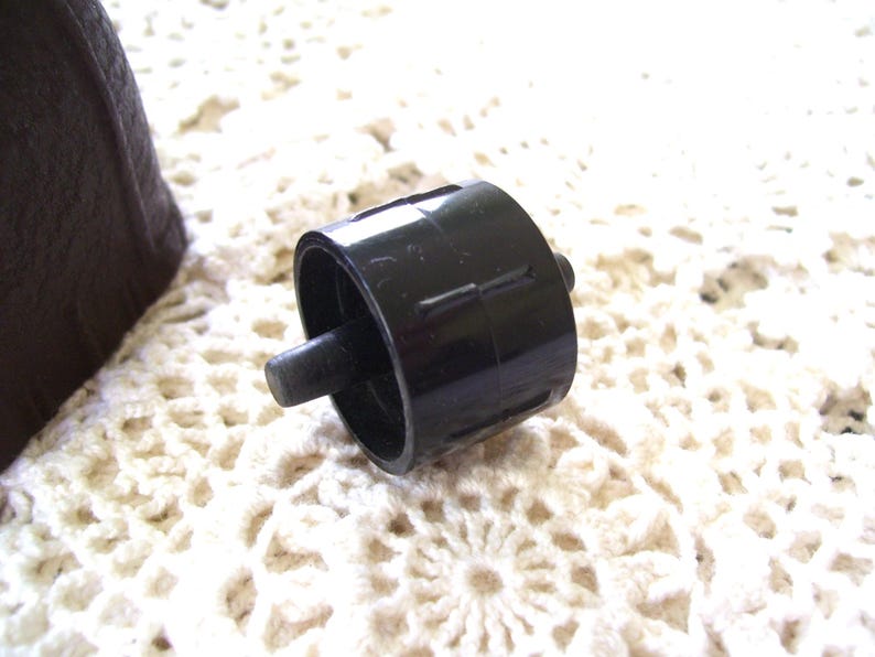 May include: A black plastic bottle stopper with a cylindrical body and a small, protruding peg. The stopper is set against a white, crocheted background. The stopper is a dark, solid color.