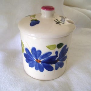 May include: A cream-colored ceramic honey pot with a matching lid featuring a pink knob. The pot is decorated with hand-painted blue flowers, green leaves, and the word Honey written in black script. A small notch is present for a spoon.