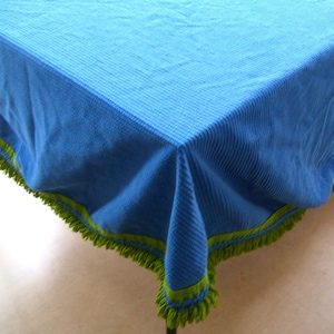 May include: A blue tablecloth with a textured surface and a green fringe trim. The tablecloth is made of a soft material and is suitable for a dining table or other surface.