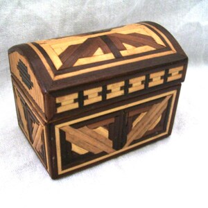 May include: A rectangular wooden box with a curved, hinged lid, featuring intricate marquetry in contrasting light tan and dark brown wood strips arranged in geometric patterns across the top, front, and side panels of the small storage chest.