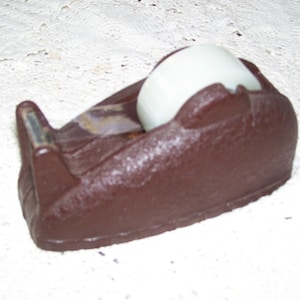May include: A brown tape dispenser with a white roll of tape. The dispenser has a textured surface and a silver cutting blade. The tape roll is partially visible, and the dispenser is sitting on a white surface.