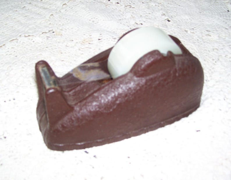 May include: A brown tape dispenser with a white roll of tape. The dispenser has a textured surface and a silver cutting blade. The tape roll is partially visible, and the dispenser is sitting on a white surface.