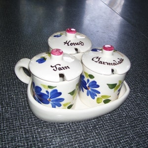 May include: A white ceramic condiment set with three lidded jars labeled "Honey", "Jam", and "Marmalade". Each jar is decorated with blue floral designs and green leaves. The set includes a matching oval tray.