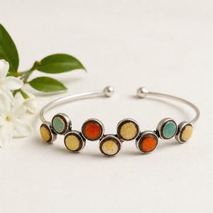 May include: A silver cuff bracelet with colourful, circular enamel accents in yellow, orange, and teal. The open-ended bracelet has small, round ball details at the ends. White flowers and green leaves are in the background.