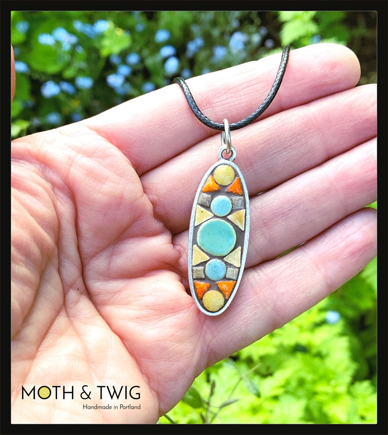 K&ouml;nnte beinhalten: Eine silberne ovale Anh&auml;nger-Halskette mit einem geometrischen Mosaik-Design. Das Mosaik besteht aus gelben, orangefarbenen, blauen und t&uuml;rkisfarbenen Fliesen. Die Halskette ist an einer schwarzen Schnur.